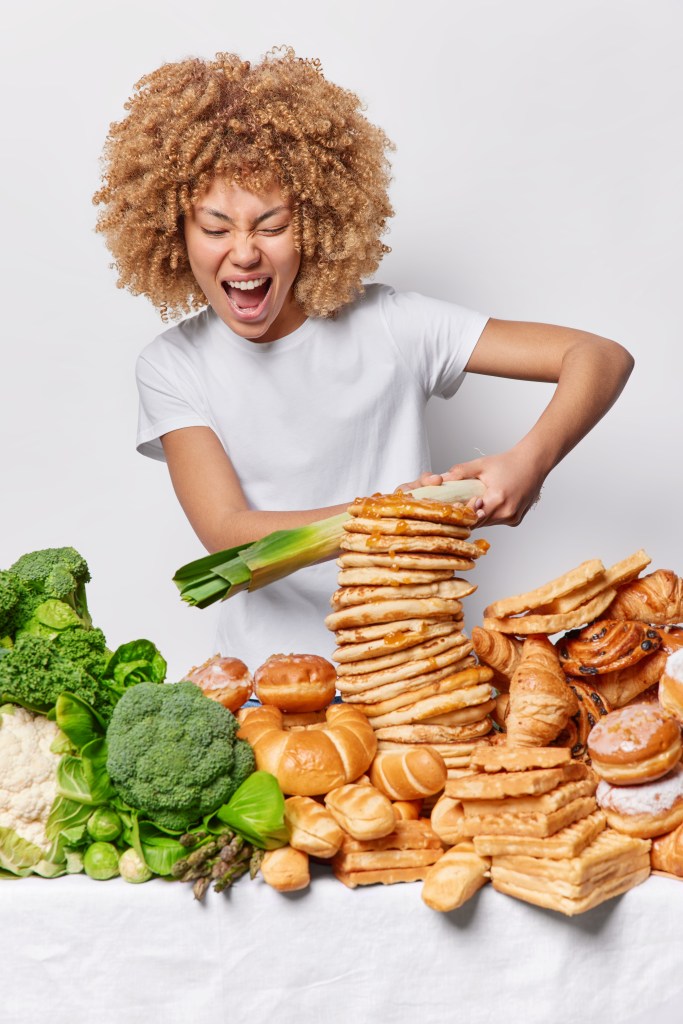 Una persona sonriente con cabello rizado sostiene una torre de pan y repostería, mientras hay vegetales como brócoli y lechuga a su lado, transmitiendo energía y emoción sobre la comida.