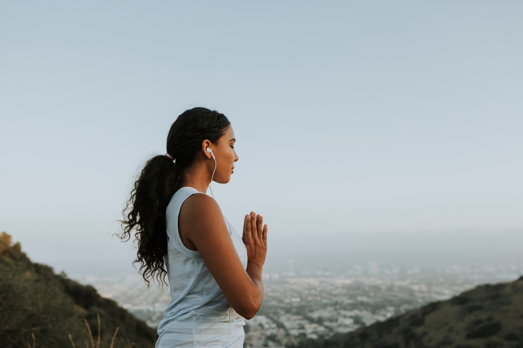 Una mujer meditando al aire libre, con vista a la ciudad, usando auriculares y con las manos en posición de oración.