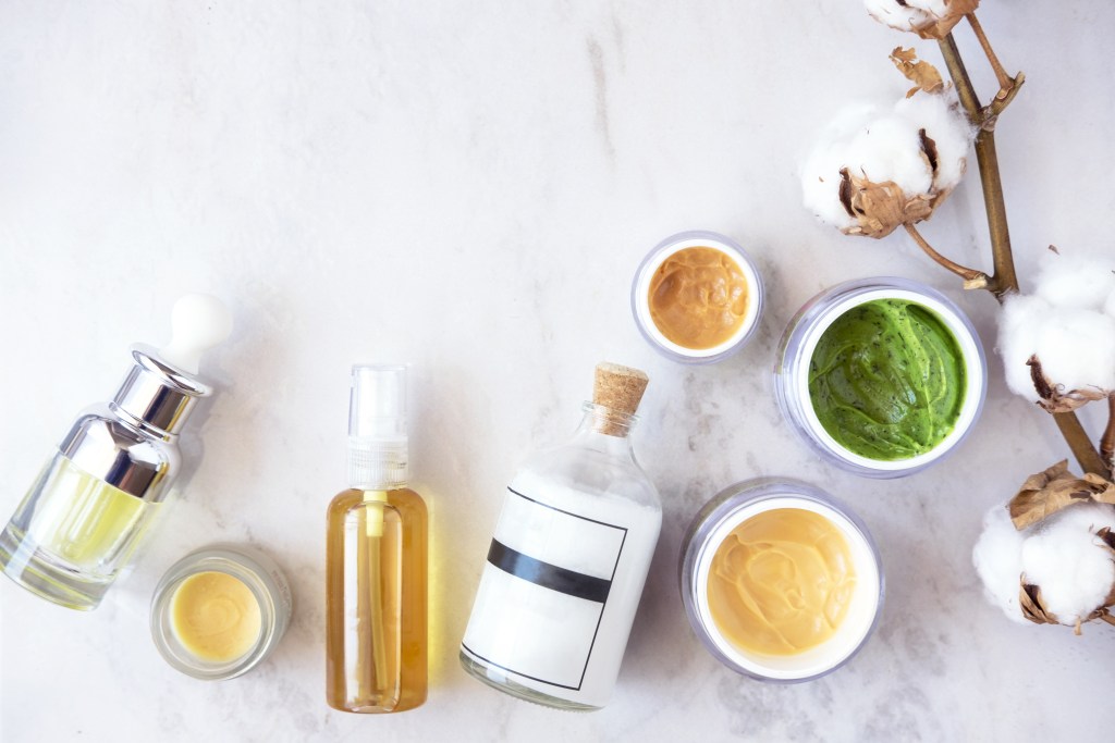 Una variedad de productos de cosmética natural sobre una superficie clara, incluyendo aceites, cremas y un ramito de algodón.
