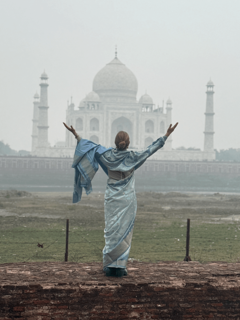Mujer de pie frente al Taj Mahal, con los brazos extendidos, vistiendo una sari azul claro, rodeada de un paisaje ligero y brumoso.