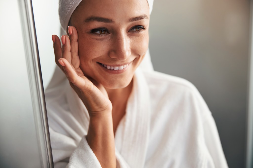Mujer sonriendo mientras se toca la cara, vestida con una bata blanca y con una toalla en la cabeza, en un ambiente luminoso.