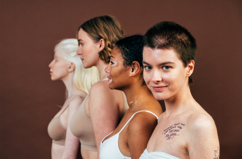 Cuatro mujeres de diferentes estilos de cabello y tonalidades de piel posan juntas, mostrando diversidad y unidad. La mujer al frente sonríe y tiene un tatuaje visible en el hombro, mientras que las demás se encuentran en perfiles, enfatizando la variedad en la belleza femenina.